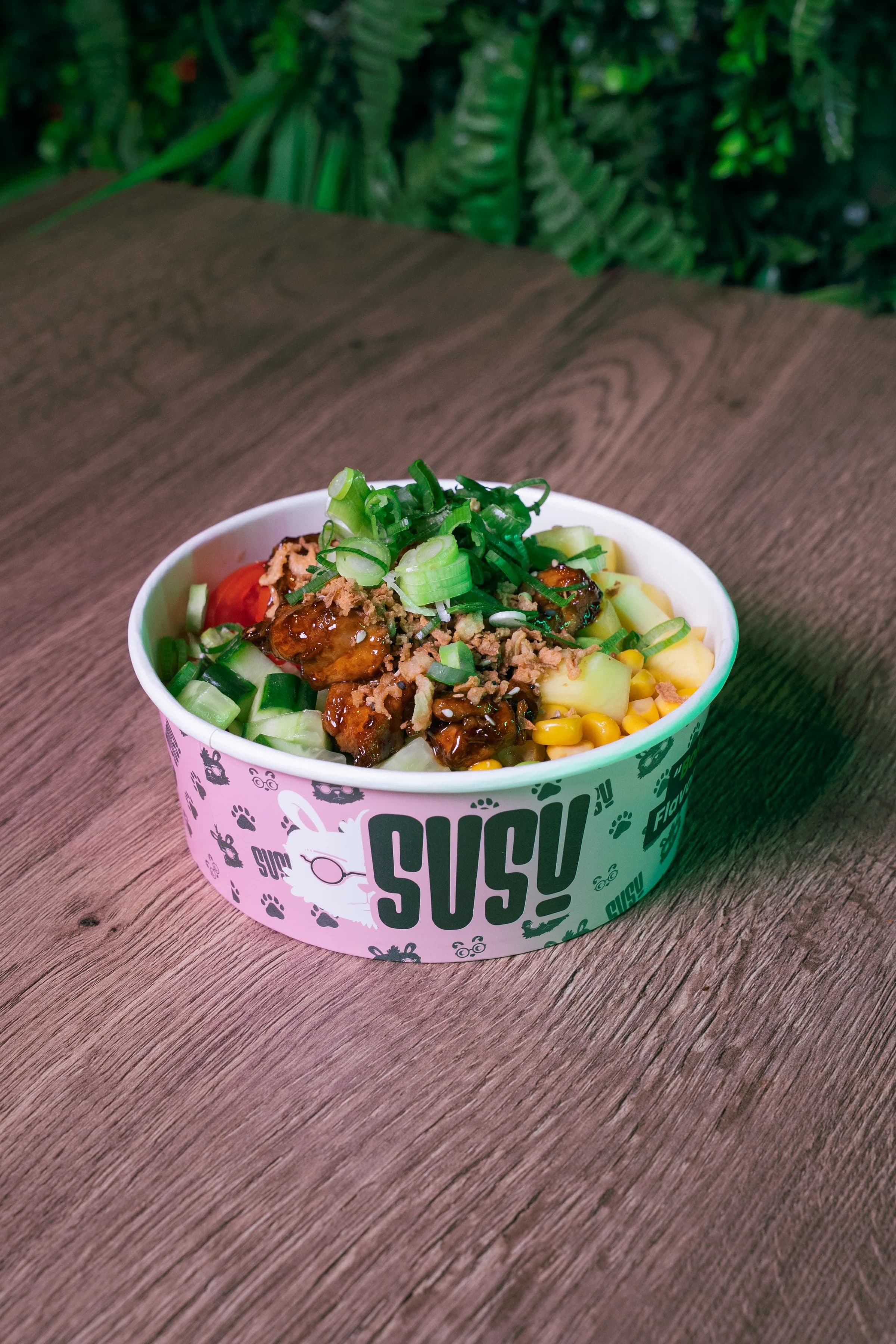 Een poké bowl met kip in een roze SUSU-bak op een houten tafel, met komkommer, tomaat en maïs