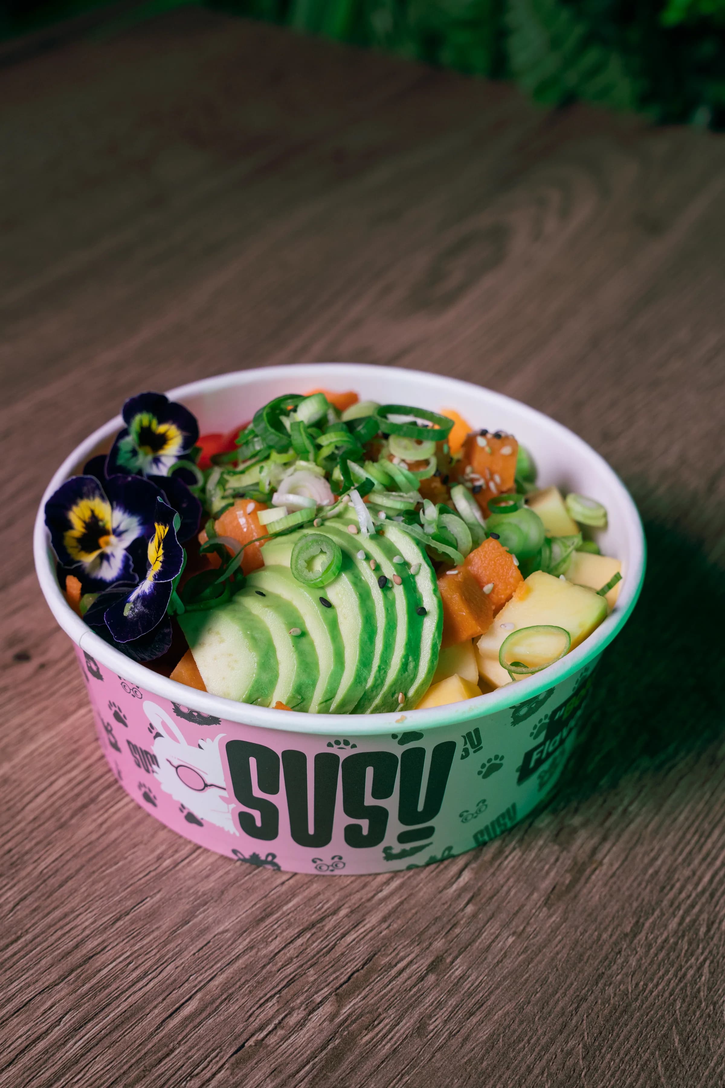 De veggie bowl in een roze SUSU-bak op een houten tafel, met avocado, zoete aardappel, ananas en viooltjes