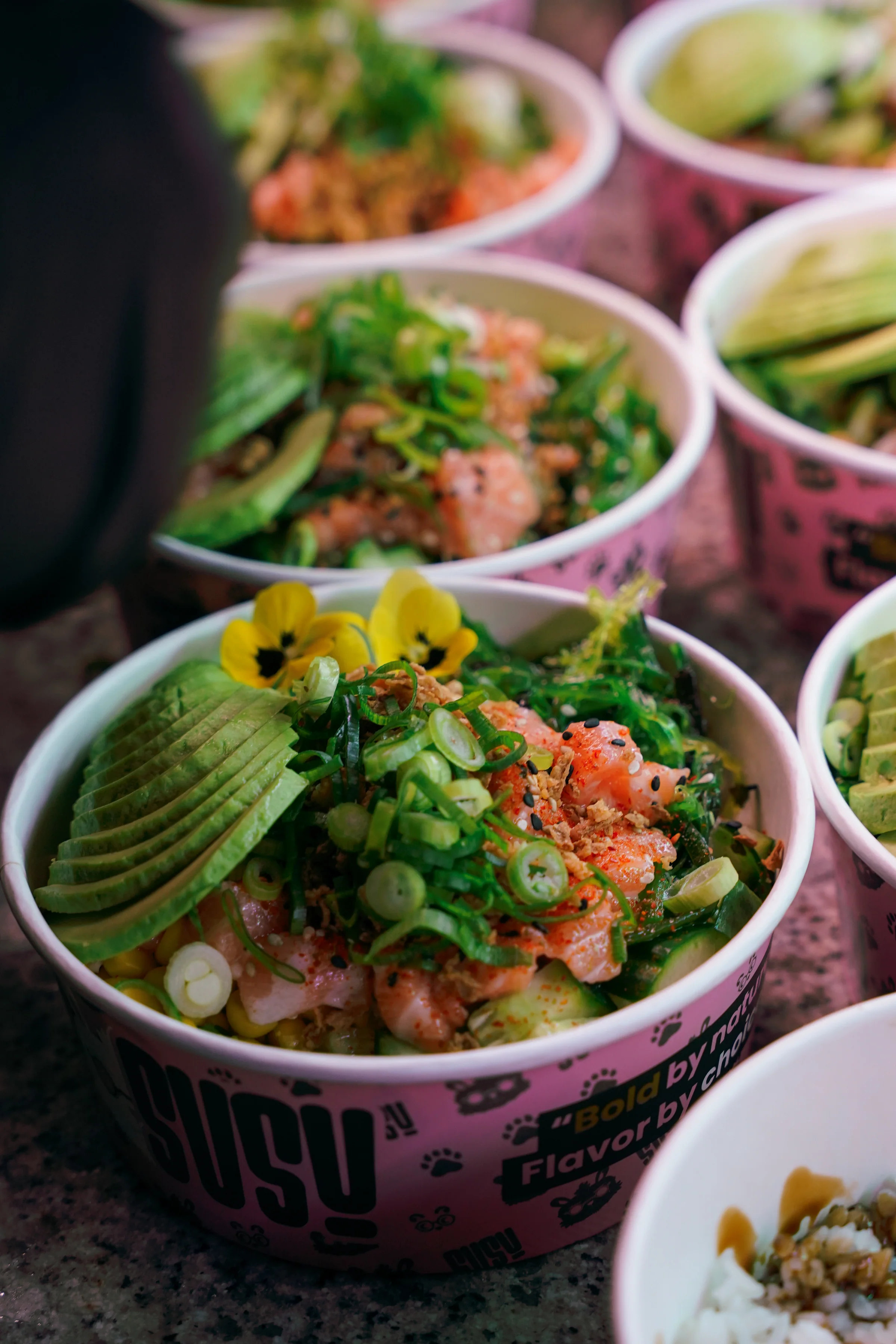 Een rij SUSU-bowls van dichtbij, met zalm, avocado, zeewier en gele viooltjes