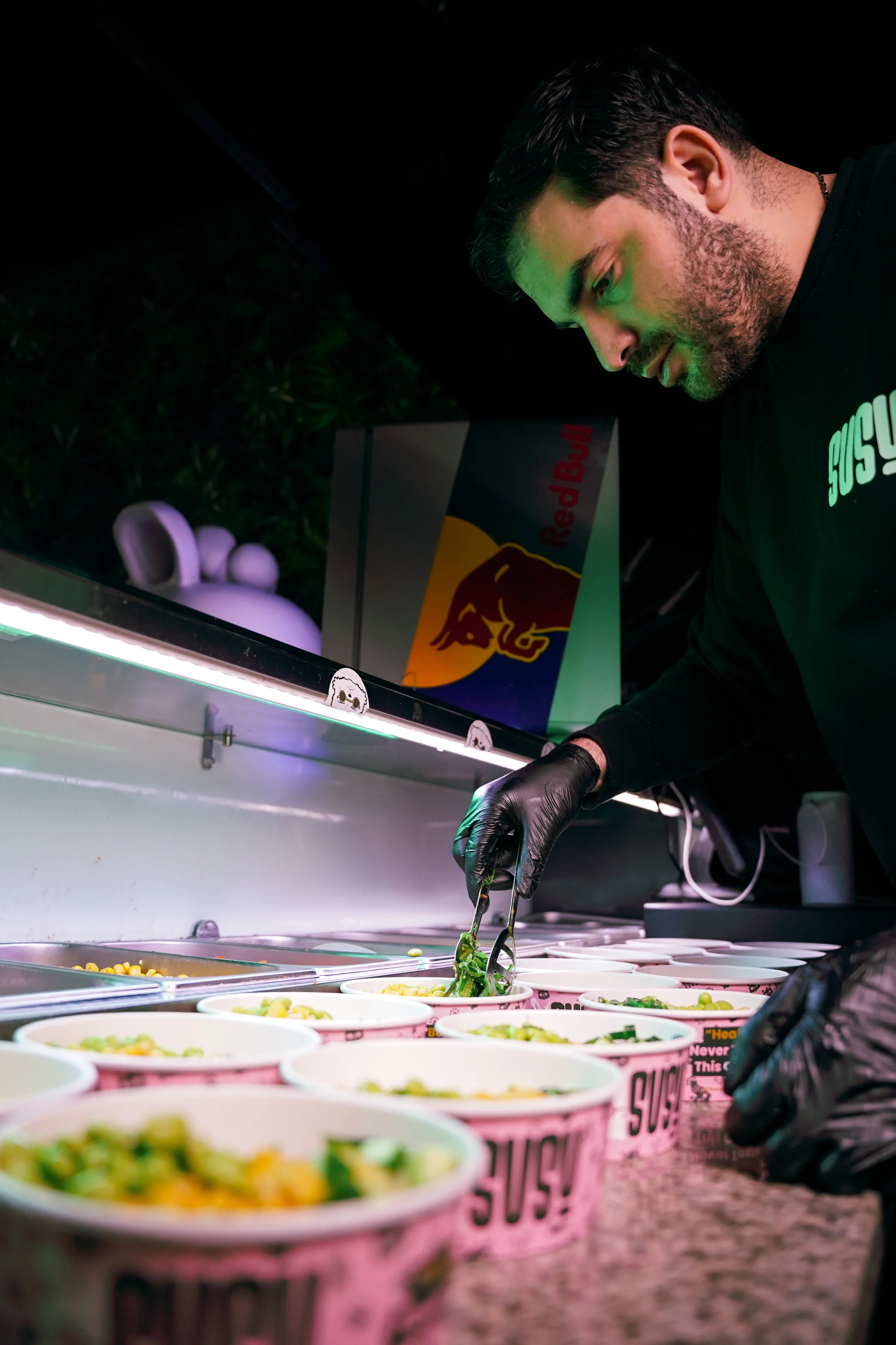 A staff member wearing gloves fills SUSU bowls at the counter, under a light box with fresh ingredients