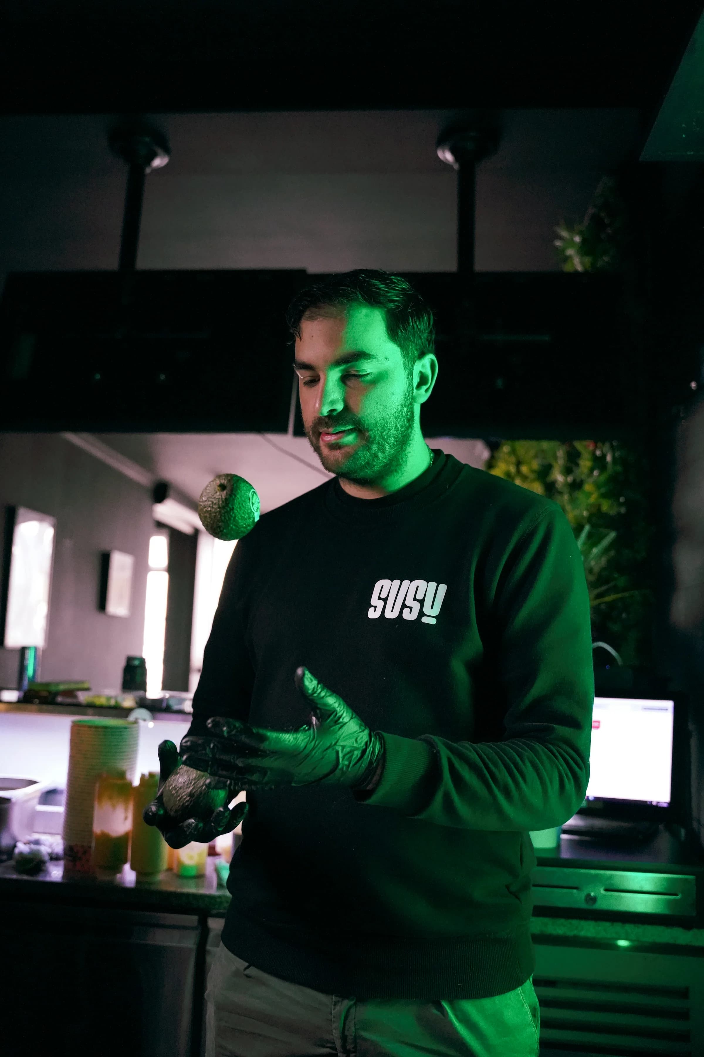 Een medewerker in een SUSU-trui en groene handschoenen houdt een avocado vast in de open keuken