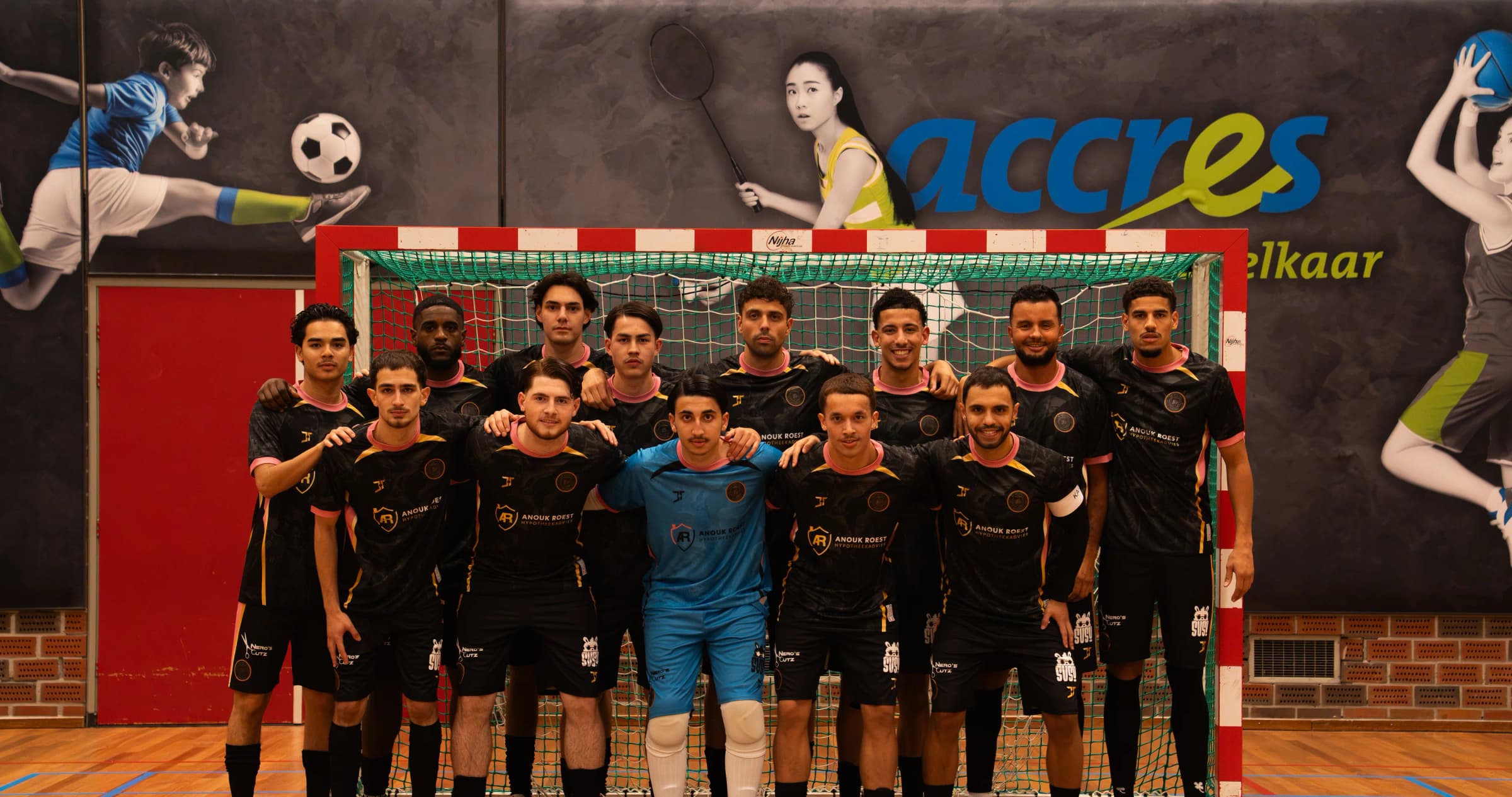 Het team in zwarte shirts met roze kragen, met een keeper in het blauw, staat voor een doel in een sporthal; het SUSU-logo staat op de broekjes